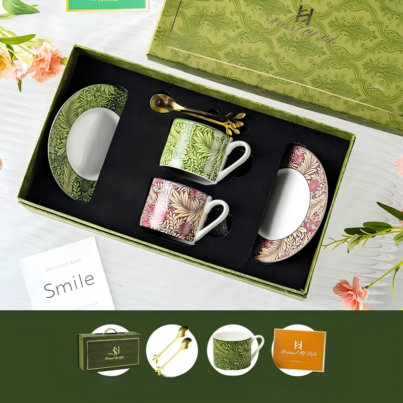 An overhead view of a luxury ceramic teacup and saucer set in a decorative green gift box, featuring two teacups and matching saucers—one in green foliage and one in pink-toned foliage—with handles and two gold spoons