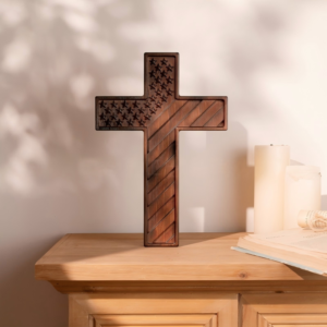 A dark-toned wooden cross carved with an American flag design featuring stars on the upper section and diagonal stripes on the rest, standing on a light wooden surface beside a book and two white candles against a softly lit background