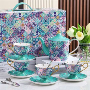 A luxury ceramic tea set arranged on a white surface, featuring a matching teapot, teacups with saucers, spoons, and a decorative gift box, all adorned with a vibrant turquoise and purple mosaic tile pattern