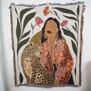 A woven throw blanket hanging against a white wall, featuring a boho-style illustration of a faceless woman in an orange patterned robe next to a seated leopard, surrounded by large dark leaves and red flowers, with fringed edges