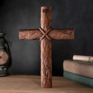 Standing rustic wooden cross made of textured log-style timber tied with a carved rope at the intersection, featuring "INRI" carved at the top and set on a wooden surface next to vintage books and a clock