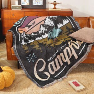 A woven throw blanket draped over a leather sofa, featuring an outdoor camping illustration with mountains, a tent, a campfire, crossed arrows, and the text "WELCOME TO OUR CAMPFIRE", finished with fringed edges