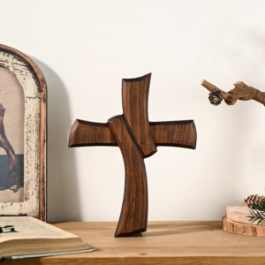 A unique, dark-stained carved wooden cross featuring a dynamic, flowing design where two curved wooden elements intersect gracefully, standing on a wooden surface next to an open book and a framed picture