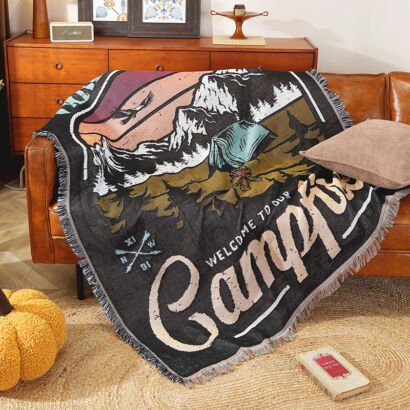 A woven throw blanket draped over a leather sofa, featuring an outdoor camping illustration with mountains, a tent, a campfire, crossed arrows, and the text "WELCOME TO OUR CAMPFIRE", finished with fringed edges