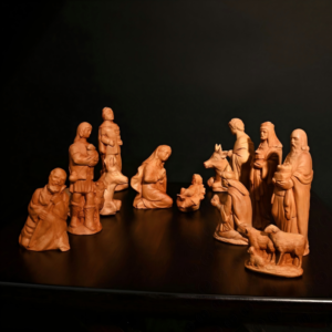 A complete set of handcarved natural wood Nativity figurines arranged on a dark surface against a black background, featuring the Holy Family, Wise Men, shepherds, animals, and the baby Jesus in a manger