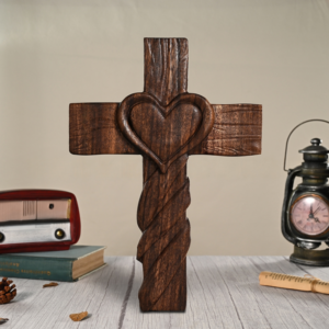 A dark-stained carved wooden cross featuring a raised heart motif at the center and a twisted rope-carved pattern along the lower vertical shaft, standing on a white wooden surface between a vintage radio and a lantern clock