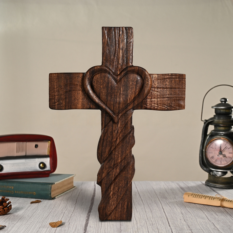 A dark-stained carved wooden cross featuring a raised heart motif at the center and a twisted rope-carved pattern along the lower vertical shaft, standing on a white wooden surface between a vintage radio and a lantern clock