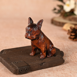 A small, dark brown wooden French Bulldog figurine sitting alertly on a textured wooden block base with a metallic corner bracket detail against a warm, softly blurred background