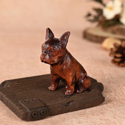 A small, dark brown wooden French Bulldog figurine sitting alertly on a textured wooden block base with a metallic corner bracket detail against a warm, softly blurred background