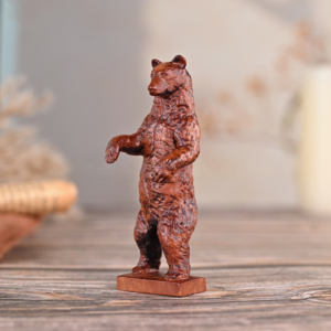 A small standing wooden bear figurine with a rich brown textured finish, posed on its hind legs on a rustic wooden surface with a soft, blurred background