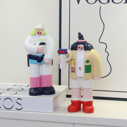 Two stylized cartoon character figurines displayed on a shelf, featuring one figure in a white outfit with pink boots holding a cup and another figure in a beige jacket with red boots holding a mug