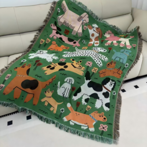 A green woven throw blanket draped over a light-colored sofa, featuring a playful all-over illustration of various stylized cartoon dogs in different breeds, colors, and poses against a green background, finished with fringed edges