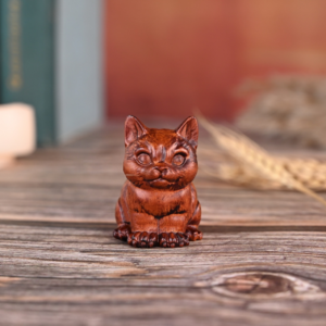 A small, handcarved wooden cat figurine with a warm reddish-brown finish, sitting upright on a rustic wooden tabletop with a blurred background