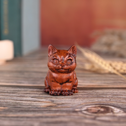 A small, handcarved wooden cat figurine with a warm reddish-brown finish, sitting upright on a rustic wooden tabletop with a blurred background