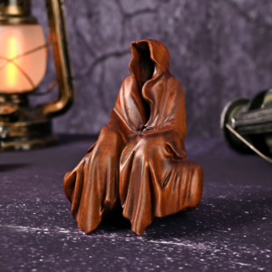 A smooth brown wood-finished figurine of a mysterious, shrouded figure wearing a deep-hooded cloak sitting on a dark surface next to a glowing vintage lantern