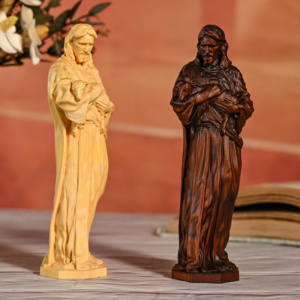 Two standing Good Shepherd Jesus statues displayed side-by-side on a wooden surface, one in a light natural wood finish and the other in a dark walnut brown finish, depicting Jesus holding a lamb