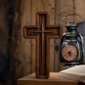 Dark stained carved wooden cross with a beveled inset border and central four-point star starburst carving, standing on a wooden surface next to an antique lantern clock and open book