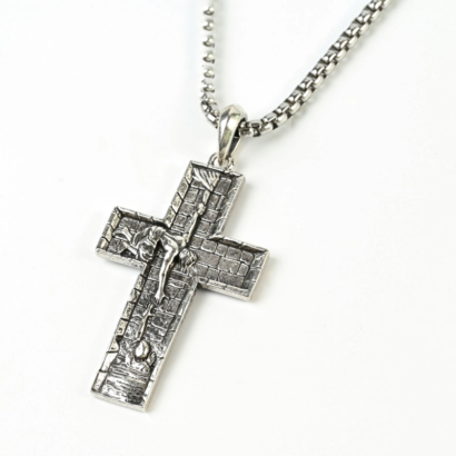 Silver-toned stainless steel cross pendant featuring an embossed relief scene set against a brick-patterned background, attached to a box chain on a plain white surface