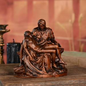 Bronze-finished statue of Michelangelo's Pieta depicting the Virgin Mary holding the body of Jesus Christ across her lap, placed on a wooden surface next to a black candle