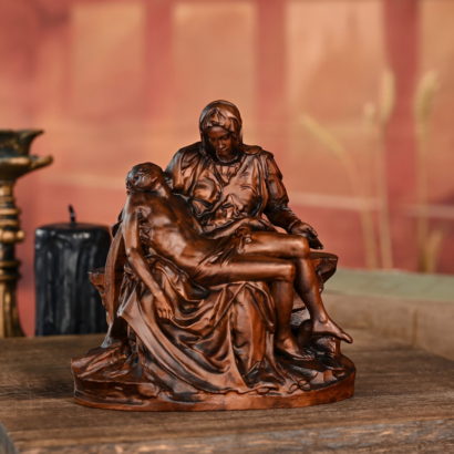 Bronze-finished statue of Michelangelo's Pieta depicting the Virgin Mary holding the body of Jesus Christ across her lap, placed on a wooden surface next to a black candle
