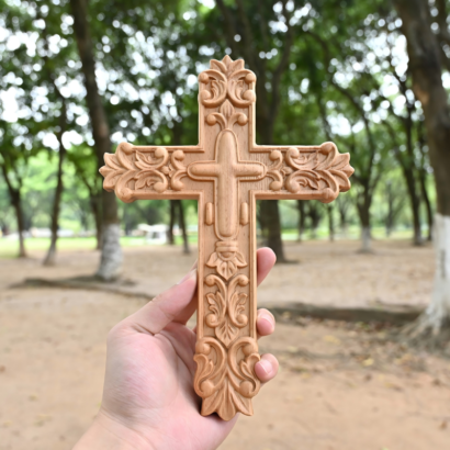 Hand holding an intricately carved natural wood cross featuring floral acanthus scrollwork and a raised central cross against a blurred park background