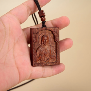 Hand holding a rectangular carved wooden pendant depicting the Sacred Heart of Jesus framed by an ornate arch, attached to an adjustable black cord string