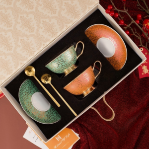 An overhead view of a luxury ceramic teacup and saucer set packaged inside a decorative gift box, featuring two teacups and matching saucers—one set in teal green and the other in orange—with gold handles, pedestal bases, and two gold spoons