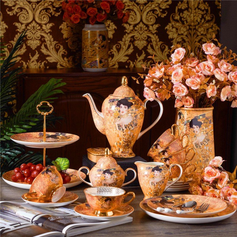 A lavish porcelain tea and dinner set featuring intricate golden motifs and classical portrait illustrations in the style of Gustav Klimt, displayed on a table alongside floral vases, a tiered fruit tray, and scattered dinnerware