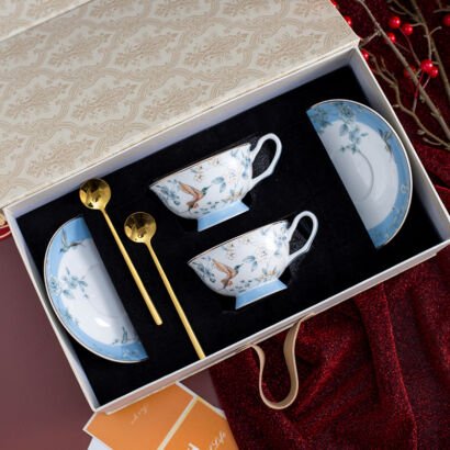 An overhead view of a luxury ceramic teacup and saucer set in a decorative gift box, featuring two light blue and white teacups and matching saucers adorned with hummingbirds and floral details, along with two gold spoons