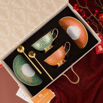 An overhead view of a luxury ceramic teacup and saucer set packaged inside a decorative gift box, featuring two teacups and matching saucers—one set in teal green and the other in orange—with gold handles, pedestal bases, and two gold spoons