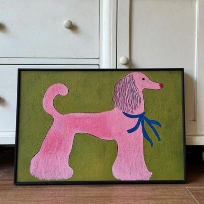 A horizontal framed canvas print resting on a wooden surface against a white cabinet, featuring a whimsical painting of a stylized pink poodle with a blue ribbon on a green background