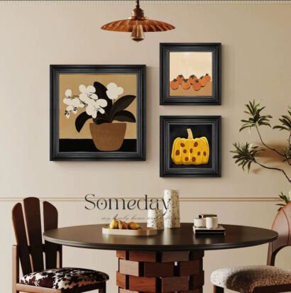A dining room wall featuring a gallery set of three black-framed art prints: a large square print of white orchids in a pot on the left, and two smaller square prints stacked vertically on the right depicting small fruits and a textured yellow pumpkin