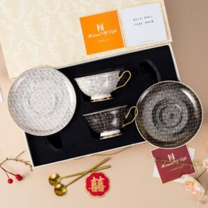 An overhead view of a luxury ceramic teacup and saucer set in a decorative box, featuring two teacups and matching saucers—one light and one dark—with an intricate woven texture pattern, gold handles, pedestal bases, and two gold spoons