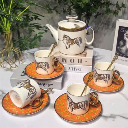 An elegant porcelain tea set displayed on a white tabletop, featuring a white teapot with a horse motif, matching teacups with gold handles, vibrant orange patterned saucers, and gold spoons