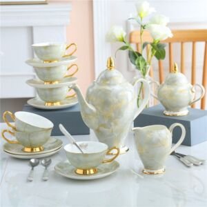Elegant white and soft grey marbled porcelain tea set displayed on a white tabletop, featuring a teapot with gold accents, milk pitcher, sugar bowl, teacups with saucers, and a stacked tower of cups and saucers
