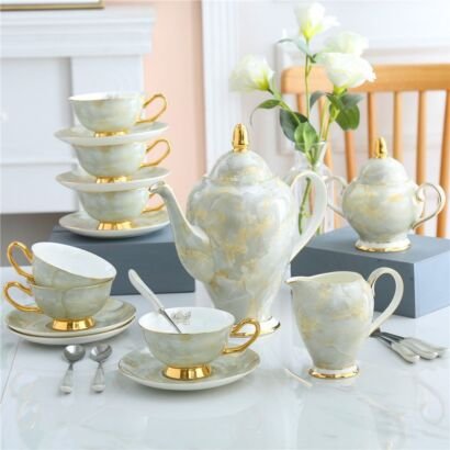 Elegant white and soft grey marbled porcelain tea set displayed on a white tabletop, featuring a teapot with gold accents, milk pitcher, sugar bowl, teacups with saucers, and a stacked tower of cups and saucers