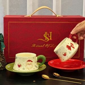 A luxury ceramic teacup and saucer set displayed in front of a red "Ritual of Life" gift box, featuring two teacup sets—one with a Santa illustration and green accents, and another with a red bow and red accents—along with matching saucers and two gold spoons