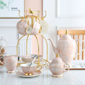 Luxury pink marbled porcelain tea service displayed on a white table, including a teapot with gold accents, milk pitcher, sugar bowl, teacups on saucers, and a gold metal bird-topped cup holder tree