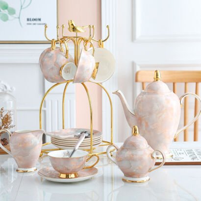 Luxury pink marbled porcelain tea service displayed on a white table, including a teapot with gold accents, milk pitcher, sugar bowl, teacups on saucers, and a gold metal bird-topped cup holder tree