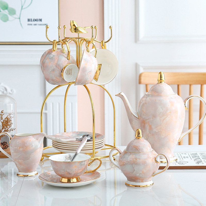 Luxury pink marbled porcelain tea service displayed on a white table, including a teapot with gold accents, milk pitcher, sugar bowl, teacups on saucers, and a gold metal bird-topped cup holder tree