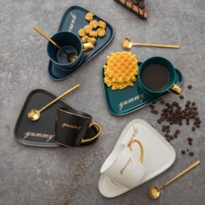 An overhead shot of a ceramic teacup and saucer set arranged on a grey surface, featuring assorted colors like dark blue, black, teal green, and white, each set with a triangular tray, gold spoons, a gold handle, and the text "yummy"
