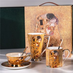 A luxury ceramic set arranged on a white surface, featuring a matching teacup with saucer, a tall mug, and a standard mug with spoons, all set against a large decorative gift box showcasing Gustav Klimt's famous painting "The Kiss" in shimmering gold tones