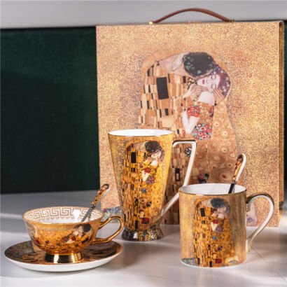 A luxury ceramic set arranged on a white surface, featuring a matching teacup with saucer, a tall mug, and a standard mug with spoons, all set against a large decorative gift box showcasing Gustav Klimt's famous painting "The Kiss" in shimmering gold tones