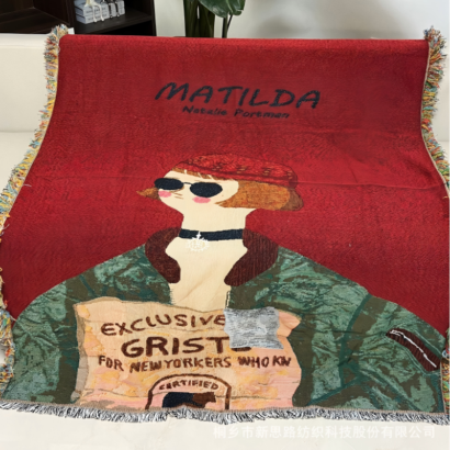 A red woven throw blanket spread out, featuring an illustrated portrait of a character in sunglasses and a green patterned jacket with text reading "MATILDA" and "EXCLUSIVE...", finished with fringed edges