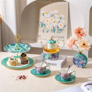 An artful tablescape featuring a Matisse-style floral art print backdrop, a glass and porcelain teapot with a warmer, two teacups with saucers, a two-tiered dessert stand with cupcakes, and a matching patterned vase with pink flowers
