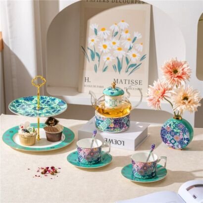 An artful tablescape featuring a Matisse-style floral art print backdrop, a glass and porcelain teapot with a warmer, two teacups with saucers, a two-tiered dessert stand with cupcakes, and a matching patterned vase with pink flowers