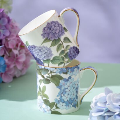 Two stacked white porcelain mugs with gold rims, one printed with purple hydrangeas and the other with light blue hydrangeas, resting on a pastel surface near fresh flowers