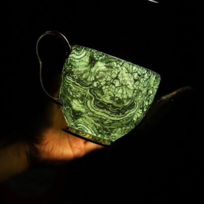Hand holding a glowing, translucent green ceramic teacup with a swirling malachite marble pattern against a dark black background