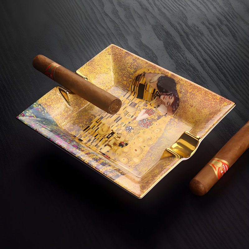 Square gold-accented ceramic cigar ashtray printed with Gustav Klimt’s famous painting The Kiss featuring dual cigar rests on a dark wood surface