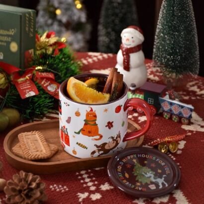 A festive holiday setup on a red patterned tablecloth, featuring a white ceramic mug filled with a hot drink, orange slices, and cinnamon sticks, with a matching lid, wooden tray holding cookies, a snowman figurine, and small Christmas trees in the background